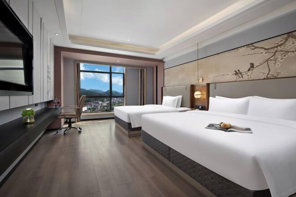 A hotel room with two beds, a desk, a chair, a large window with a city view, and a wall-mounted TV.