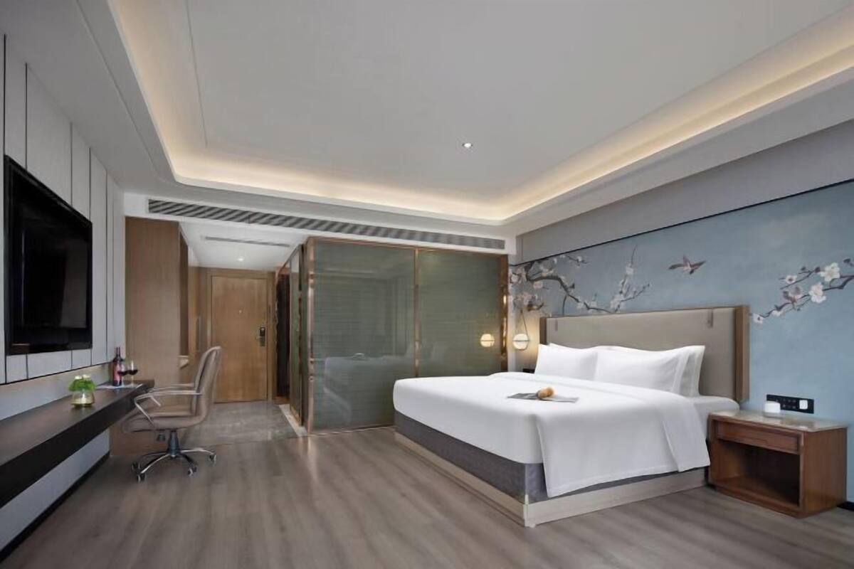 A modern hotel room with a large bed, a desk, a TV, and a glass shower enclosure.