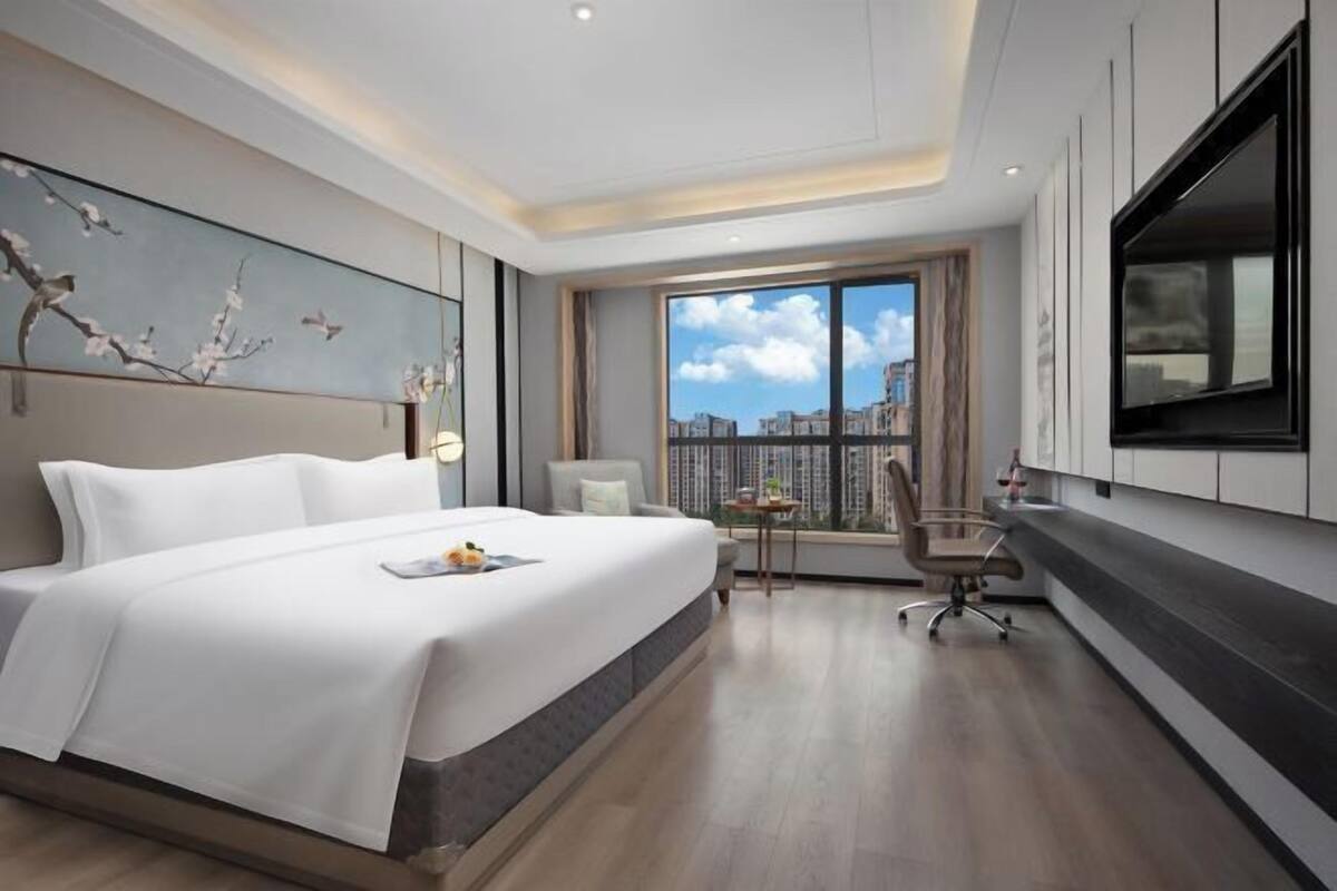 A modern hotel room with a large bed, a desk, a chair, a TV, and a city view.