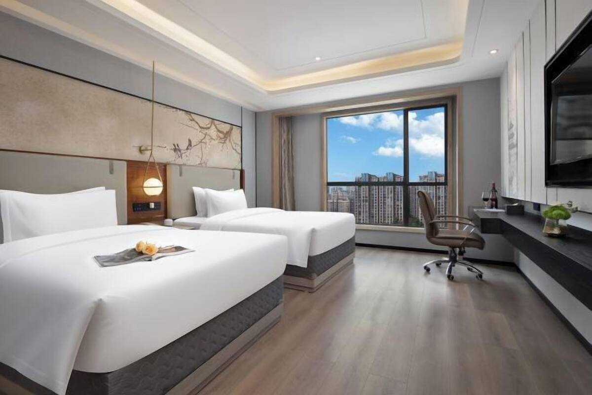 A hotel room with two beds, a desk, a chair, a large window with a city view, and a mounted TV.