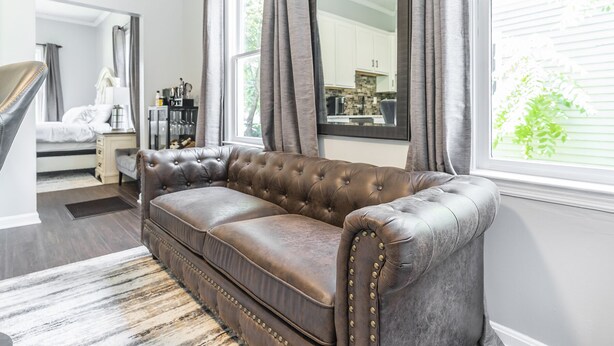 Een bruine leren bank in een woonkamer met een groot raam, een spiegel en uitzicht op een keuken.