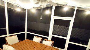 A covered patio area with a wooden table and chairs, large windows, and a ceiling fan.
