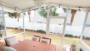 A covered patio with a wooden table and chairs, a ceiling fan, and a view of a backyard with a white fence and plants.