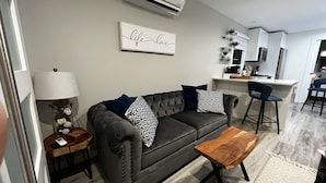 A modern living room with a grey sofa, patterned cushions, a wooden coffee table, and a kitchen area in the background.