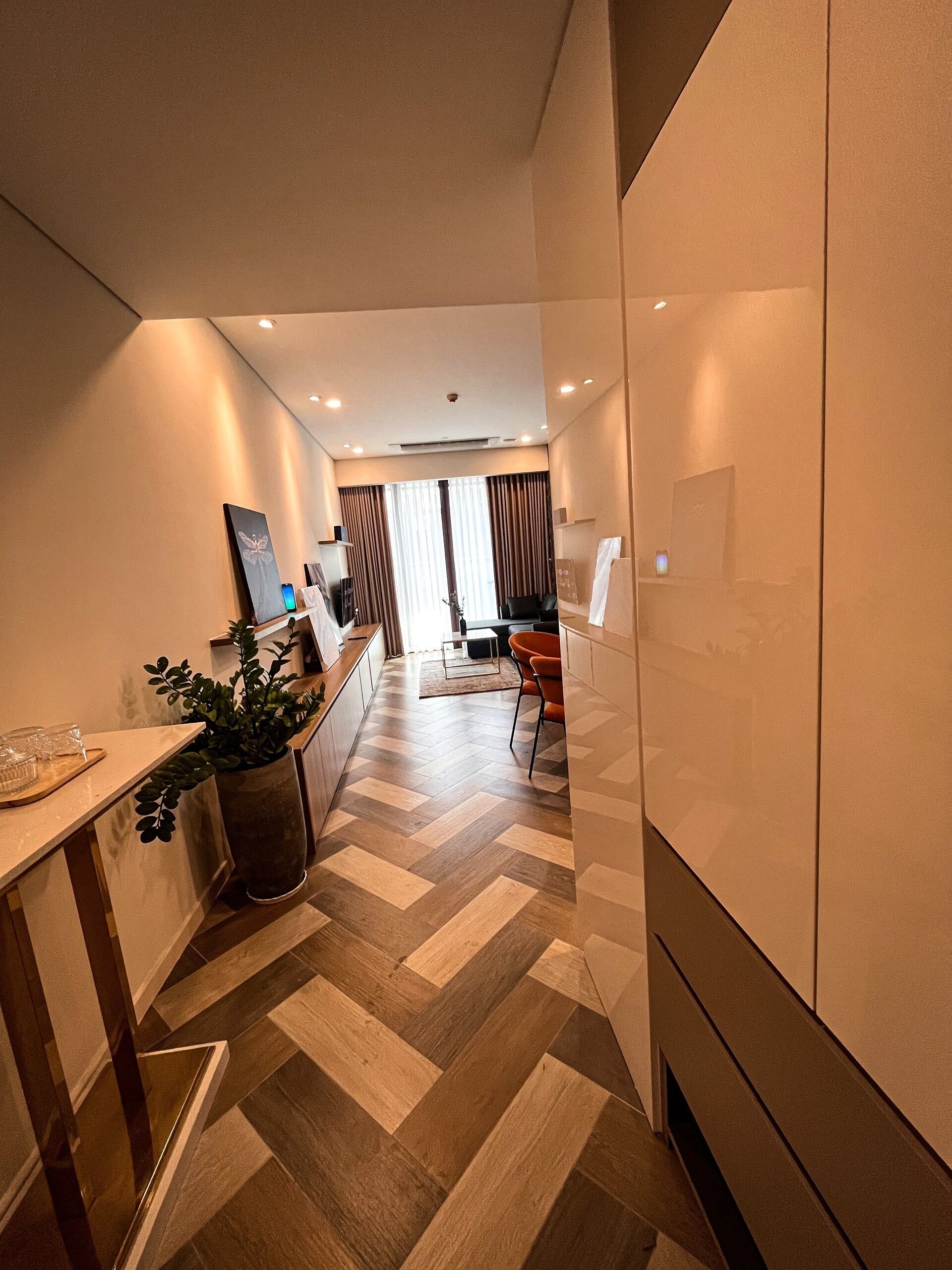 A modern hotel room with a herringbone floor pattern, a dining area with a table and chairs, and a large potted plant.