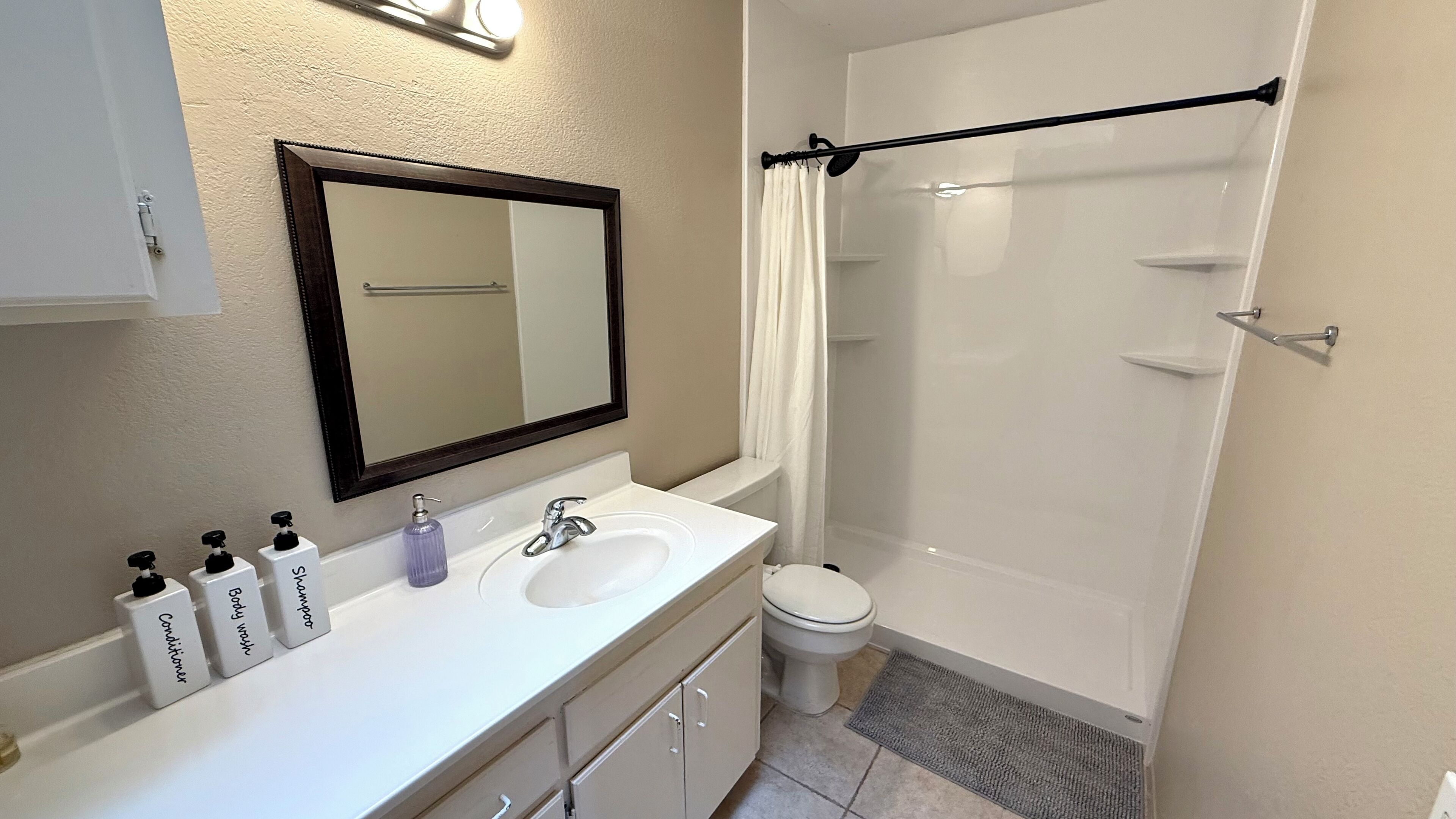 Een badkamer met een wit wastafel, spiegel en toilet. Er staan drie flessen shampoo en conditioner op de rand. Een douchegordijn is zichtbaar, en een handdoekhouder is aan de muur bevestigd.