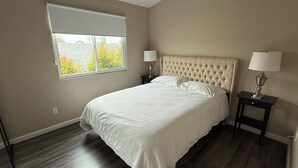A bedroom with a large bed, two bedside tables with lamps, a window with blinds, and a view of trees outside.