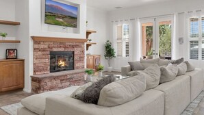 A living room with a fireplace, a large flat-screen TV mounted above it, a beige sofa, and a window with white shutters. - Natural Springs-2 acre, Games+karaoke, fit,views. (Riverside County)