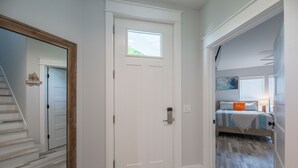 Ein Flur mit einer weißen Tür, einem spiegelnden Wandbild mit Holzrahmen und einer Treppe, die zu einem Schlafzimmer mit Bett und Fenster führt.