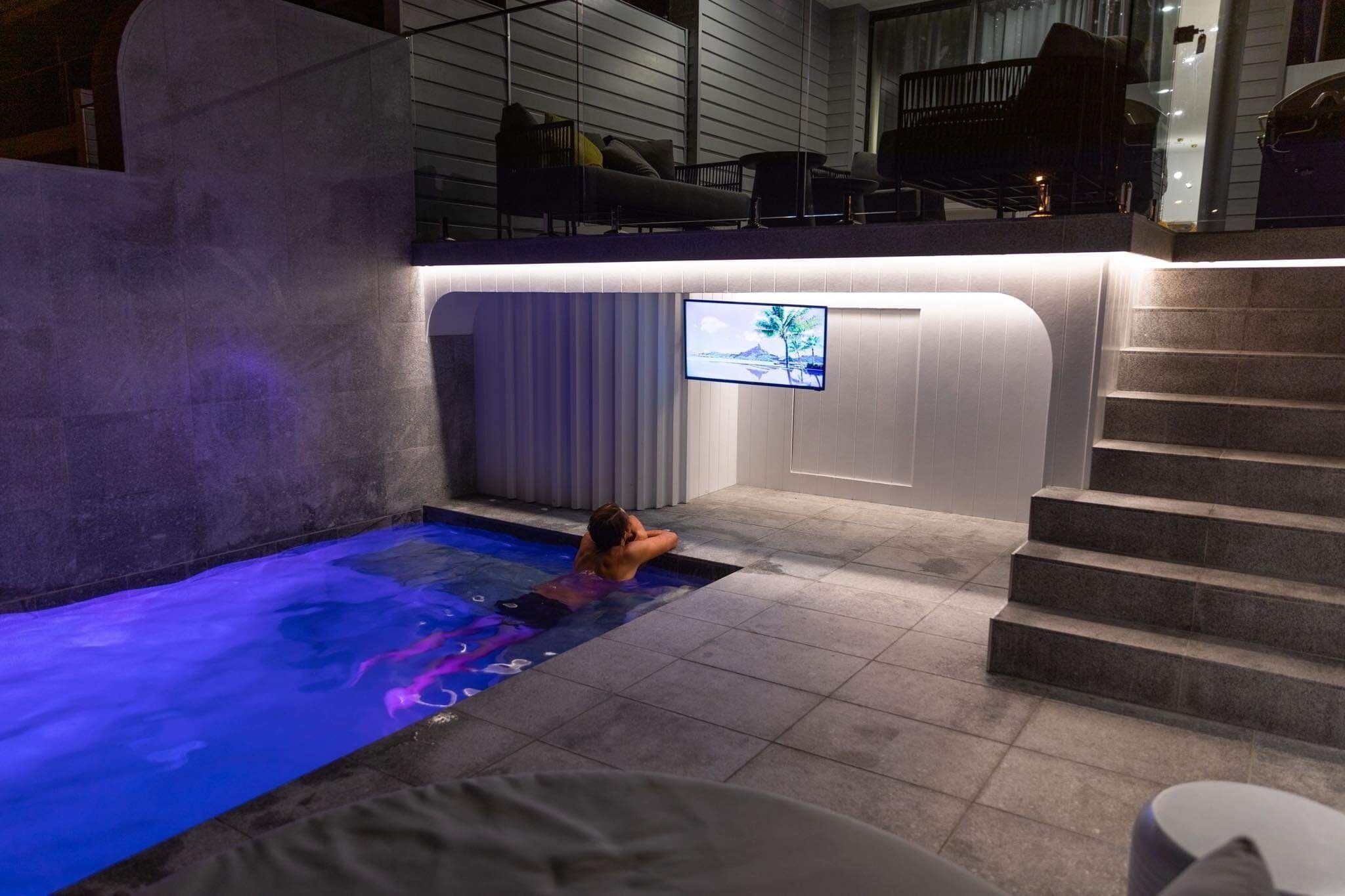 A modern indoor pool area with a person relaxing in the water, a television mounted on the wall, and a staircase leading to an upper level.