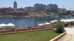 A waterfront view with a grassy area, lounge chairs, and umbrellas, overlooking a marina with boats and a cityscape in the background. - Apartment With Pool Near Beach In St Julians (Swieqi)
