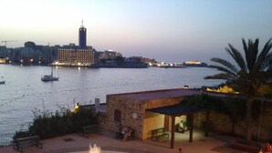 A waterfront view with a fountain, a stone building, and a distant cityscape. - Apartment With Pool Near Beach In St Julians (Swieqi)