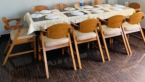 Uma área de refeições com uma mesa posta para quatro pessoas, rodeada por cadeiras de madeira com assentos acolchoados. Um ventilador de teto é visível acima da mesa.