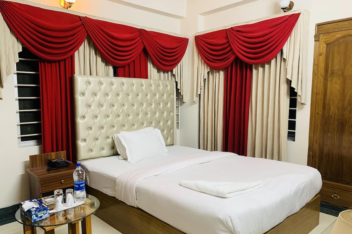 A hotel room with a large bed, red curtains, a wooden headboard, a bedside table with a phone and toiletries, and a wooden wardrobe.
