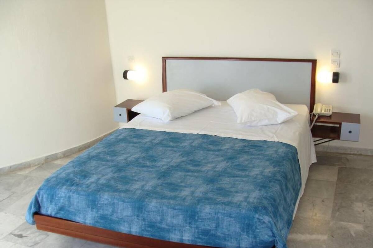 Ein Bett mit hölzernem Kopfteil, weißen Kissen und einer blauen Decke. Auf der einen Seite befinden sich Nachttische mit Lampen und einem Telefon.