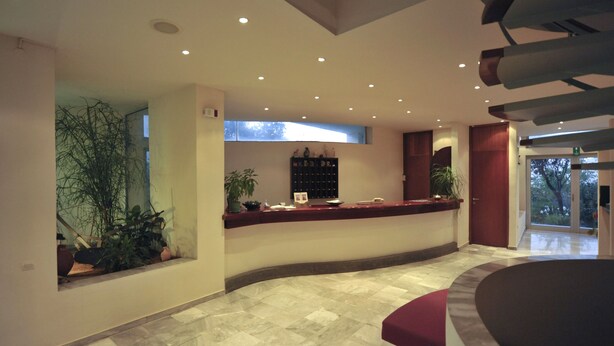A modern reception area with a curved reception desk, potted plants, and a large window providing natural light. - Rodon Hotel (Chania)