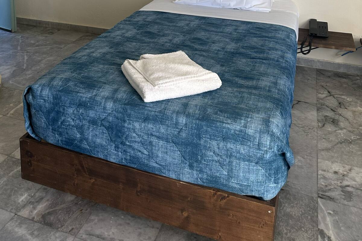 Ein Einzelbett mit blauer Decke und weißen Kissen, ein Nachttisch mit Telefon sowie ein an der Wand befestigtes Stromverteilergehäuse.
