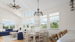 A spacious living room with a blue sofa, a dining table with chairs, and a large window offering a view of greenery. - Beachwalk Luxury Home: Heated Pool & Free Bikes (Palm Coast)