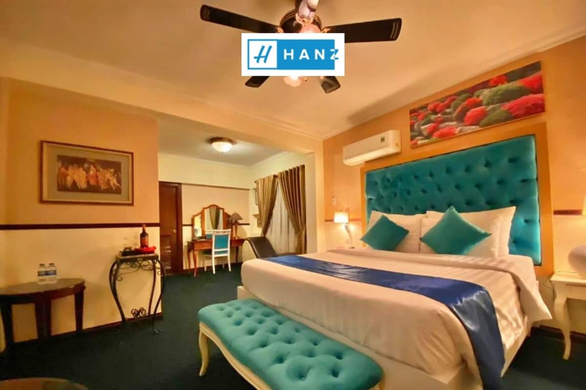 Quarto de hotel com uma cama grande, um banquinho azul estofado, uma escrivaninha com cadeira e um ventilador de teto.