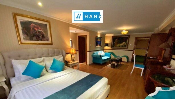 A hotel room with a large bed, a sofa, a desk, and a chair. There is a painting on the wall and a lamp on the desk. - S79 Vuon Saigon Hotel & Spa (Ho Chi Minh City)