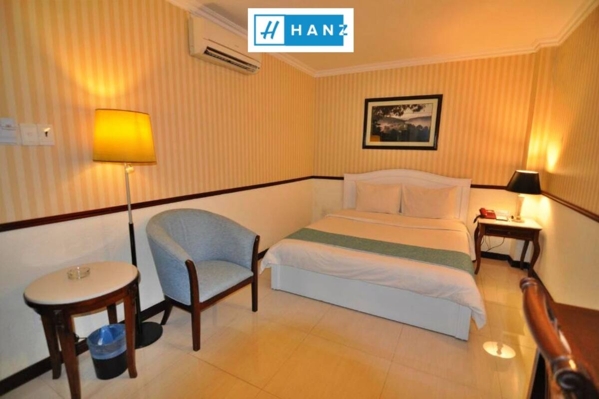 Quarto de hotel com uma cama, uma cadeira, uma mesinha e uma mesa de cabeceira com abajur.
