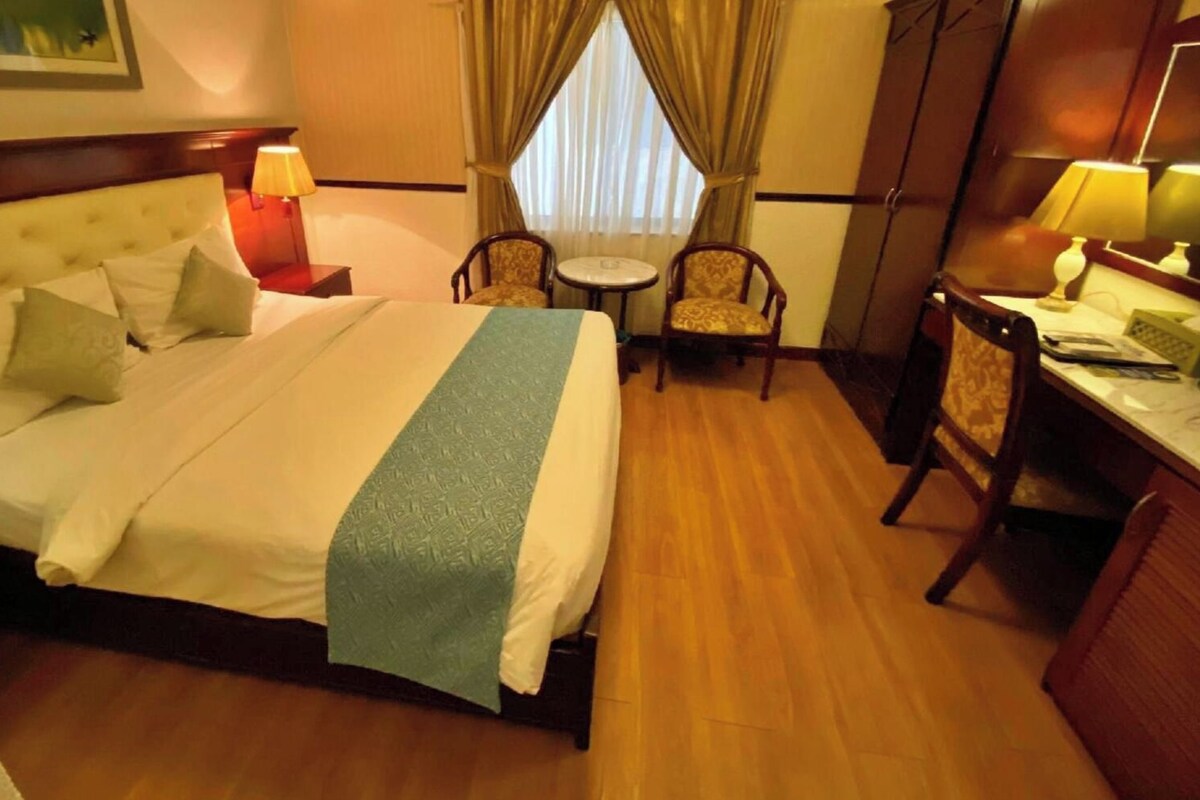 A hotel room with a large bed, a desk, two chairs, a small table, and a window with curtains.