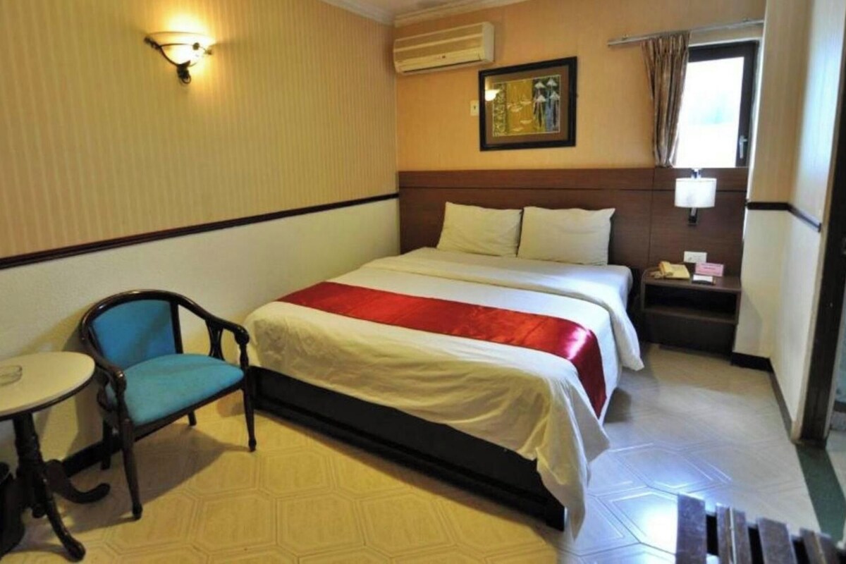 A hotel room with a bed, a chair, a small table, a nightstand, a window with curtains, and an air conditioning unit.