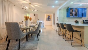 A modern dining area with a long table and chairs, a kitchen with blue cabinets, and a painting on the wall. - Elegant 4BR Home w/ Private Pool (Santiago De los caballeros)