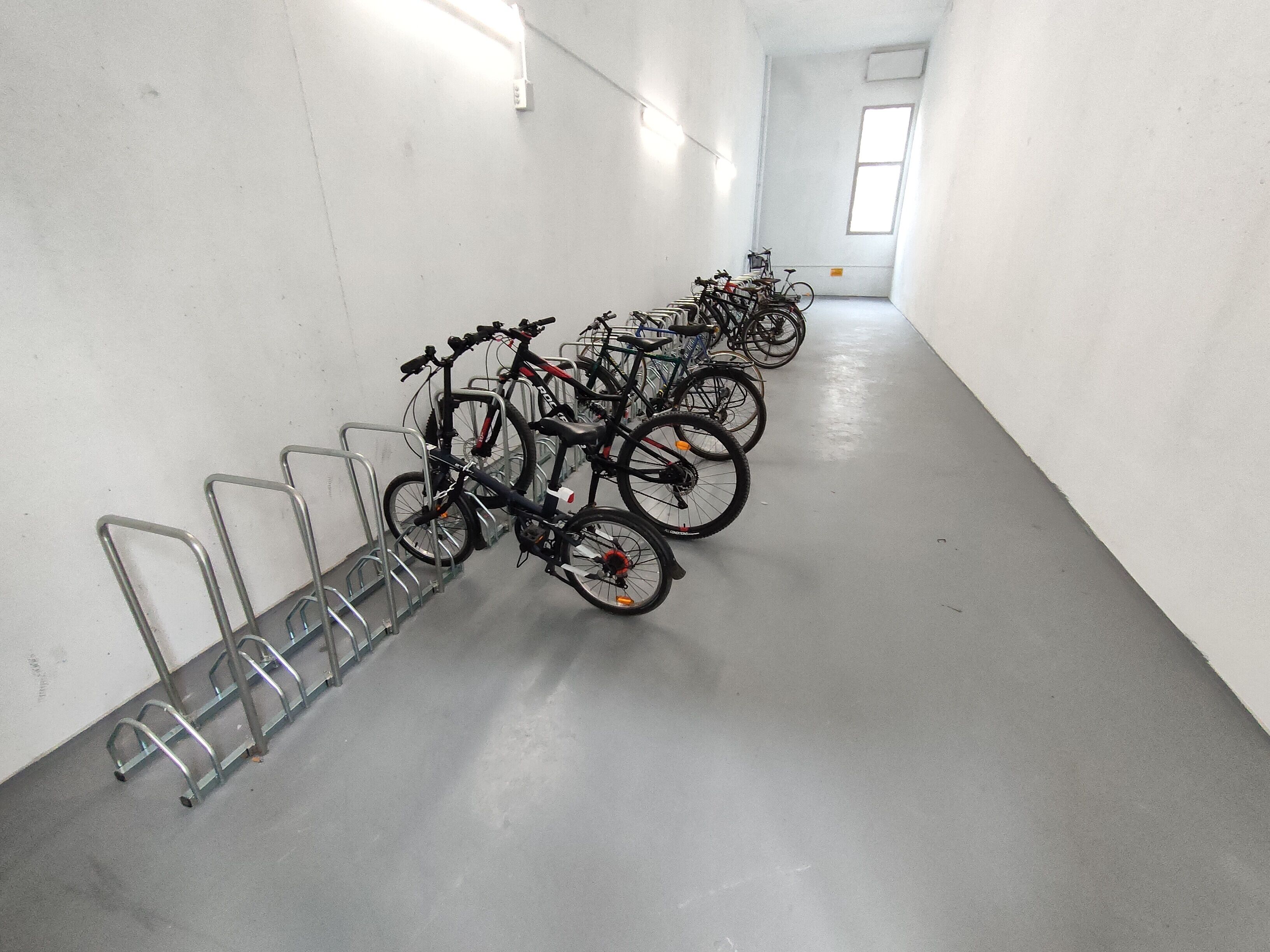 A row of bicycles parked in a well-lit, narrow hallway with white walls and a grey floor.