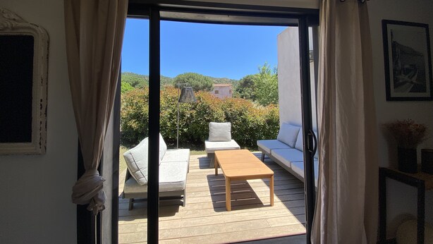 An open sliding door leading to an outdoor deck with a sofa, a table, and a view of a hillside with buildings. - Villa Olivia 10 minutes from Palombaggia without traffic jams (Porto-Vecchio)
