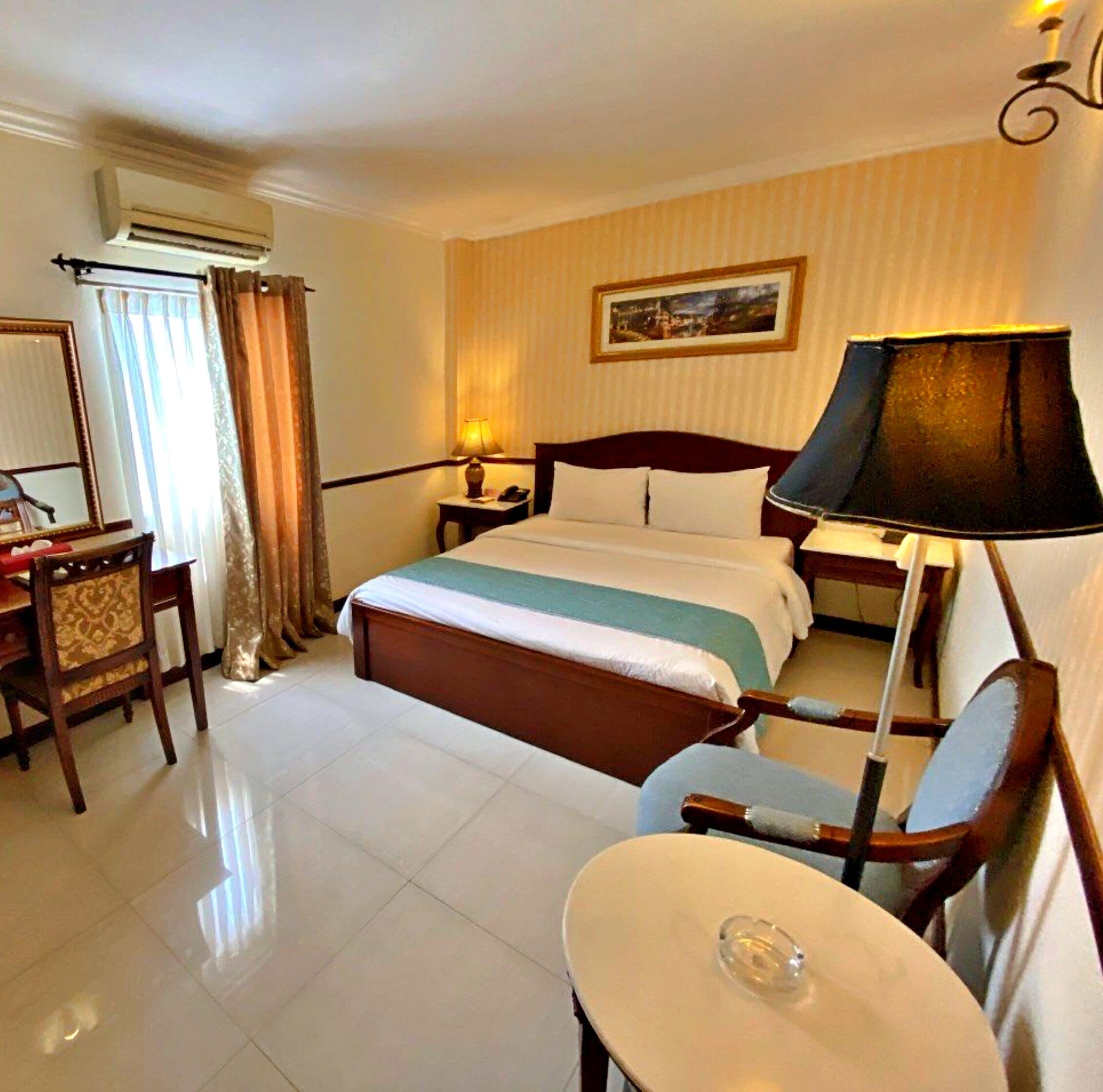 A hotel room with a bed, a desk, a chair, a table, a lamp, and an air conditioning unit.