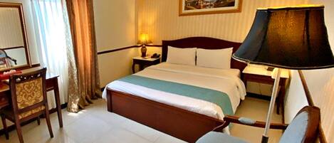 A hotel room with a bed, a desk, a chair, a table, a lamp, and an air conditioning unit.
