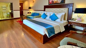 A hotel room with a large bed, a wooden headboard, a painting above the bed, and a small bedside table with a lamp.