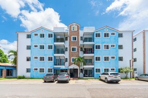 Un immeuble à plusieurs étages doté d’une entrée centrale, de balcons et d’un espace de stationnement où sont garées des voitures.