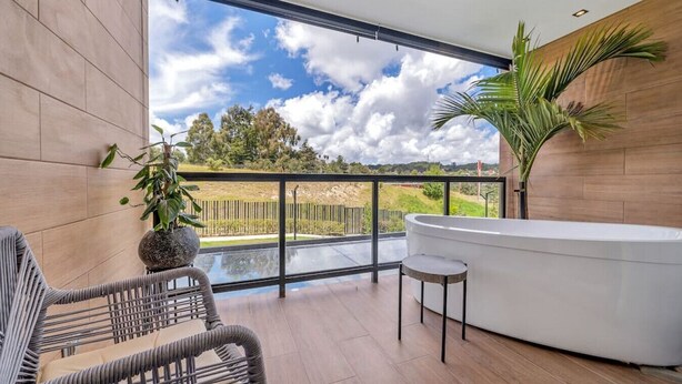 Un balcon moderne avec une porte vitrée, une baignoire blanche, des meubles en osier et une vue sur un paysage verdoyant.
