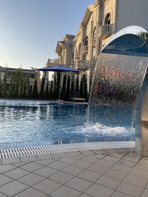 Una piscina con fuente, rodeada por una solana con baldosas y un edificio de arquitectura clásica.
