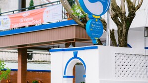 A hotel entrance with a blue and white sign, a covered outdoor seating area, and a visible US Vietnam Talent International School sign in the background. - Hoi An Nguyen Homestay (Da Nang)