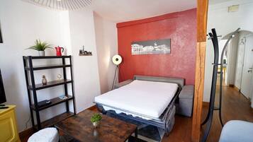 Une pièce de taille modeste comprenant un lit, une petite table, une étagère avec des objets décoratifs, une lampe sur pied et un mur d’accent de couleur rouge.