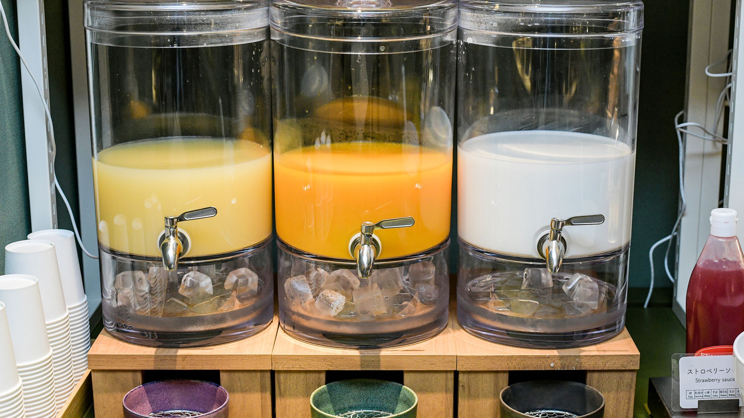 Three beverage dispensers with taps, ice, and a sign indicating a strawberry sauce option.