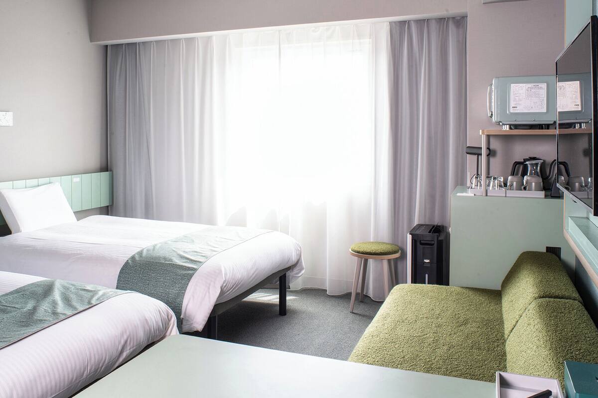 Habitación de hotel con dos camas, un escritorio, una silla, un taburete pequeño, una cafetera y un televisor.