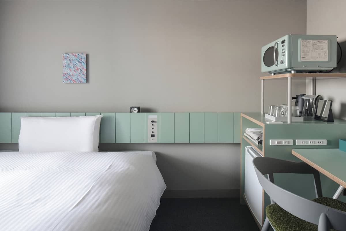 Habitación de hotel con cama, escritorio, silla, microondas y un pequeño estante con diversos objetos.