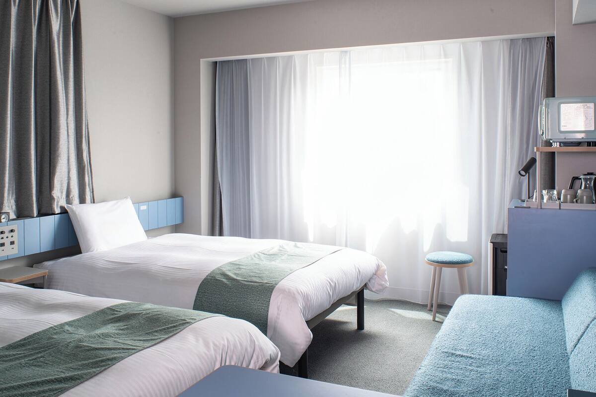 Habitación de hotel con dos camas, un escritorio, una silla, un taburete pequeño, un televisor y una ventana con cortinas.