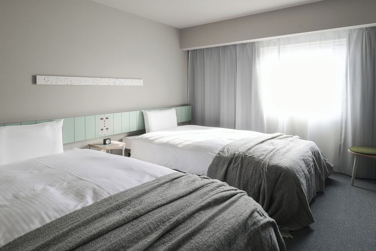 Una habitación de hotel con dos camas, una mesita de noche, una pequeña mesa y un ventanal con cortinas.