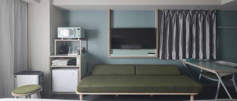 Ein kleines Hotelzimmer mit einem Bett, einem Schreibtisch, einem Fernseher, einer Minikühlschrank und einem Regal mit diversen Gegenständen.