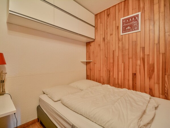 A bedroom with a wooden headboard, a bed with white bedding, a bedside table with a lamp, and a small shelf.