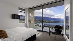 Un dormitorio con cama, televisor y una silla con vistas a montañas y un lago.