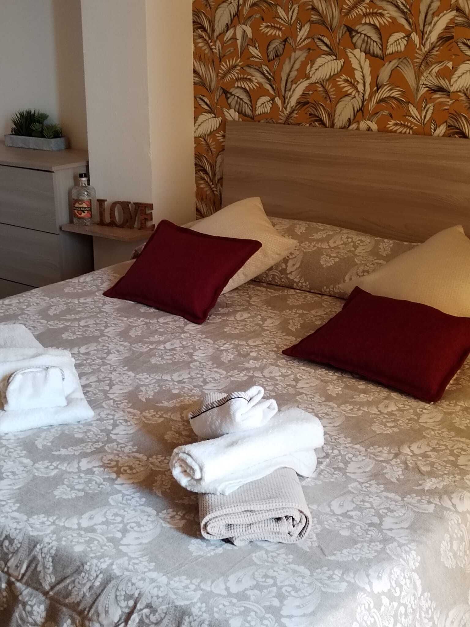 A neatly made bed with floral-patterned bedding, two red pillows, and rolled towels.