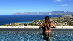 A woman in a pool overlooking a coastal landscape with mountains in the distance.