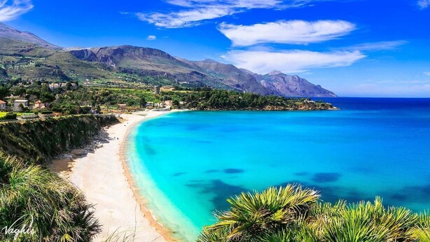 A coastal landscape with a beach, clear turquoise waters, and mountainous terrain in the background.