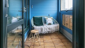 Une pièce bleue avec une chaise en osier, un petit tabouret et une fenêtre donnant sur des immeubles.